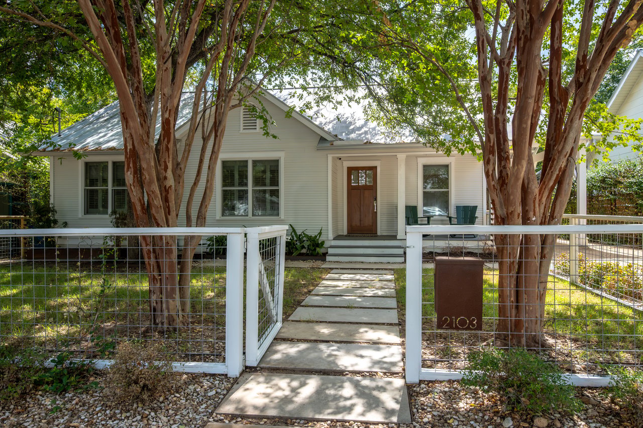 Single-story light-colored house behind an open white fence, shaded by mature trees.