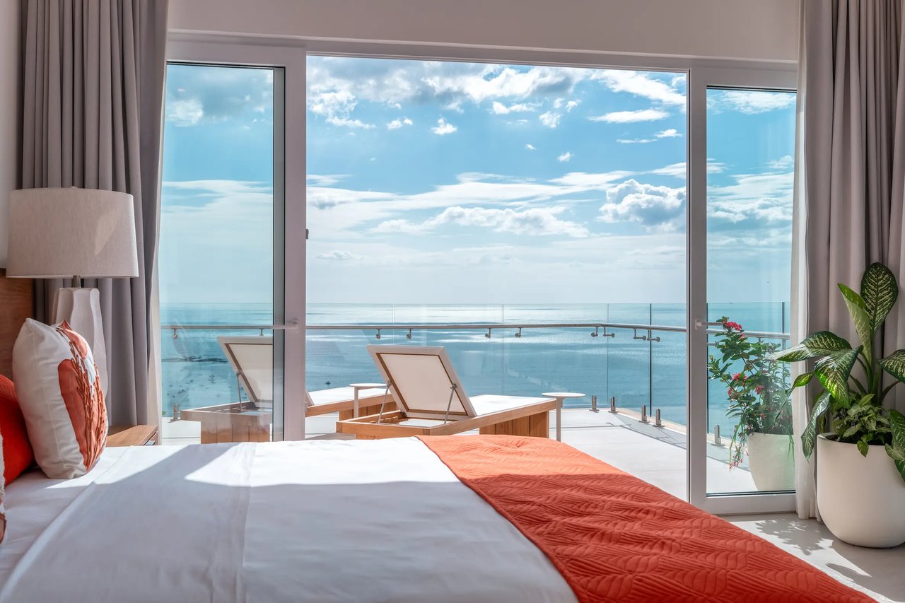 Condo bedroom with coral bed runner and floor-to-ceiling doors opening to an ocean-view balcony.