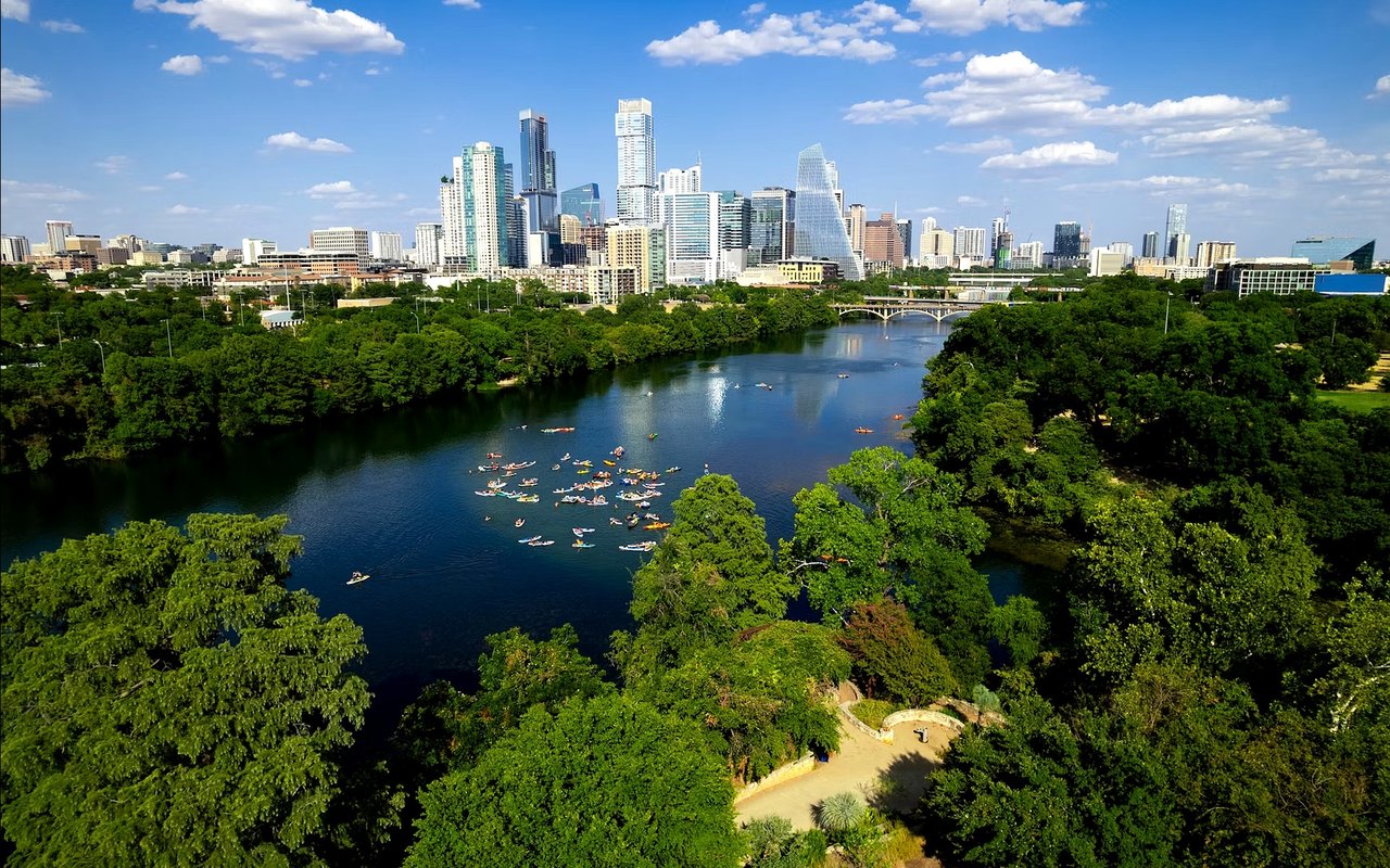 Downtown Austin skyline above Lady Bird Lake, with kayaks on the water and green parkland along the banks.
