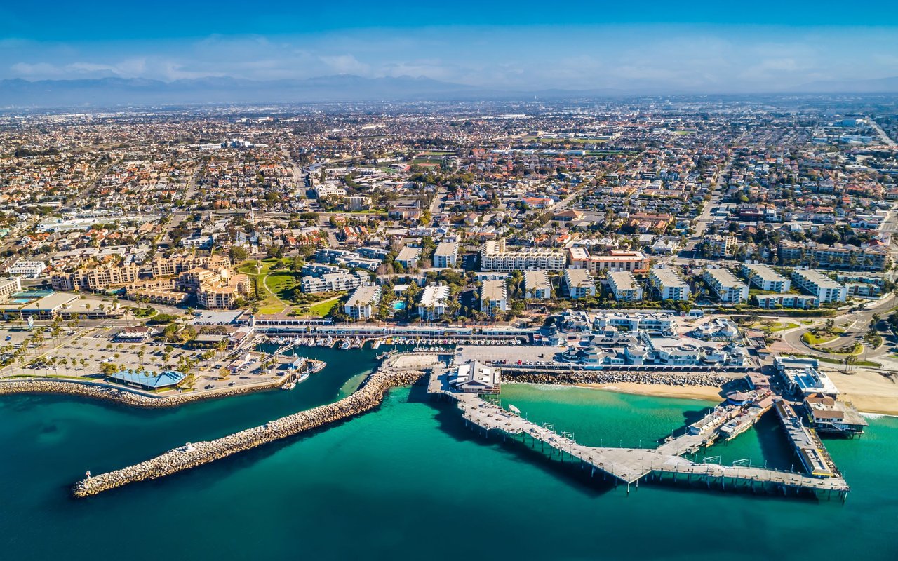 Coastal marina and breakwaters in front of a sprawling neighborhood, with mountains on the horizon.