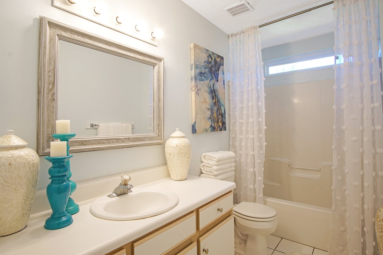 Bright bathroom with light blue walls, featuring a large mirror above a white sink and countertop. Two blue candle holders and decorative vases are present. A patterned shower curtain and abstract wall art add elegance.Bright bathroom with light blue walls, featuring a large mirror above a white sink and countertop. Two blue candle holders and decorative vases are present. A patterned shower curtain and abstract wall art add elegance.