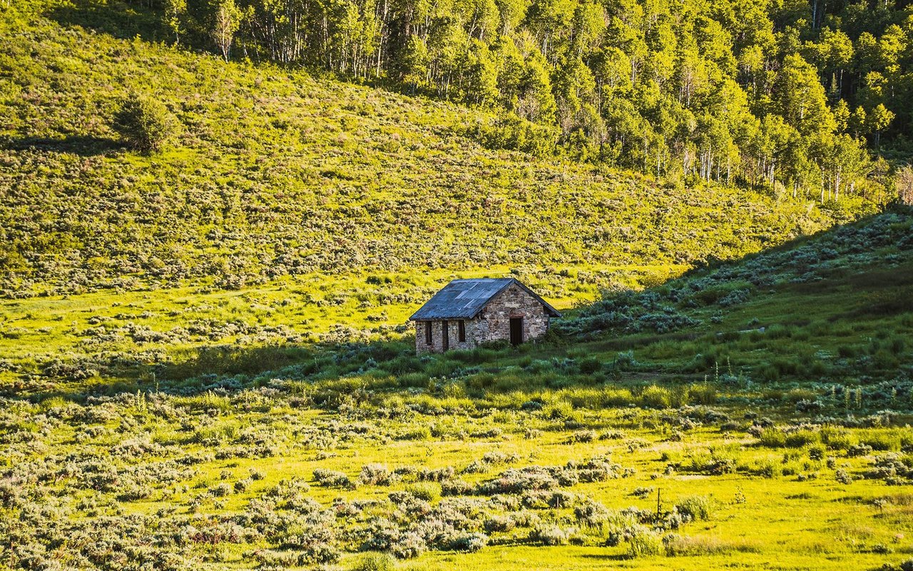 Small stone cabin in a meadow beneath a sunlit hillside of golden aspens.