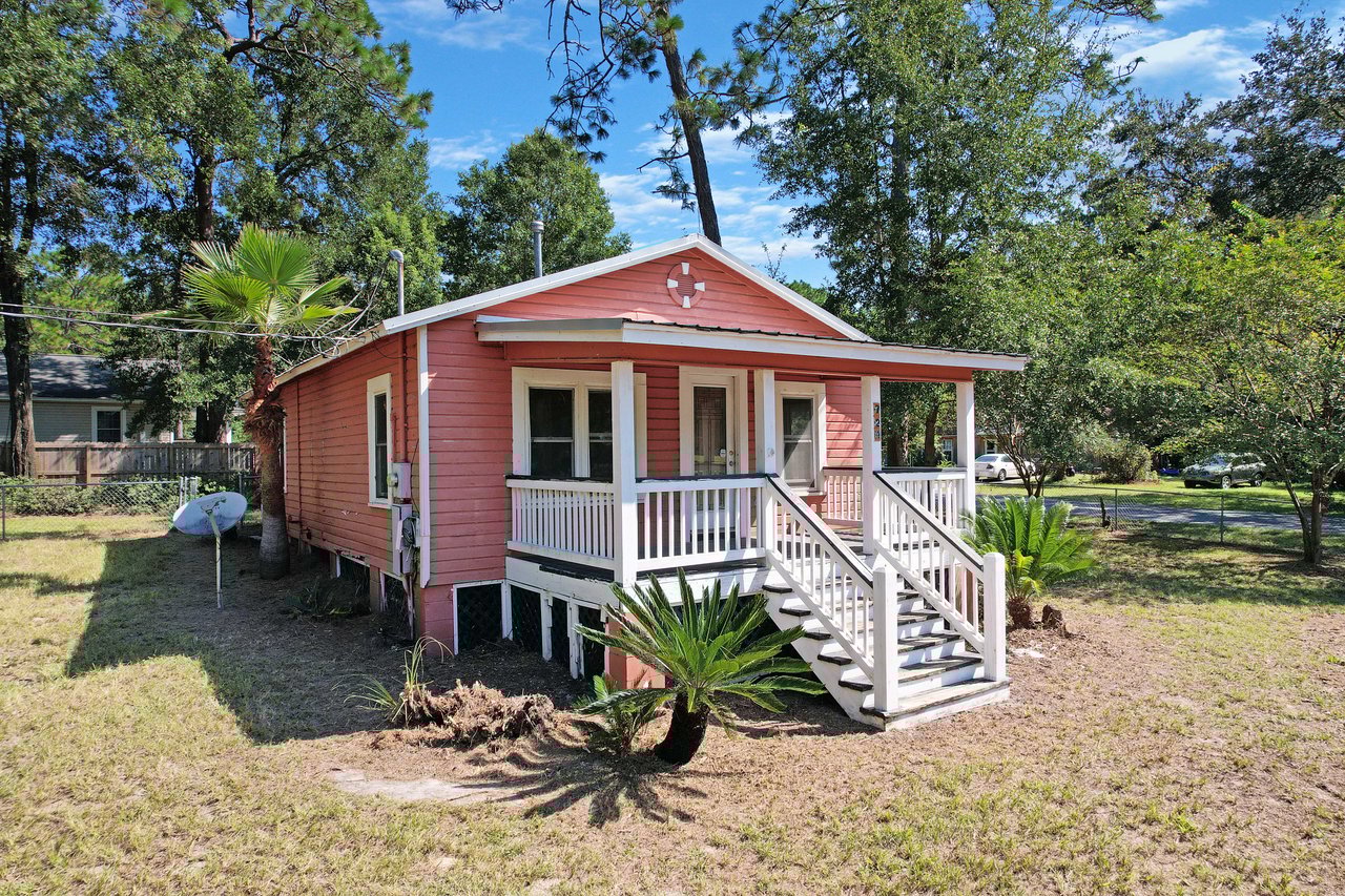 Front exterior of a small pink cottage-style home with white trim and a covered front porch. The house features white railings, steps leading to the entrance, and is surrounded by a grassy yard with palm plants and mature trees in the background. 724 flagg street, tallahassee, florida 32305