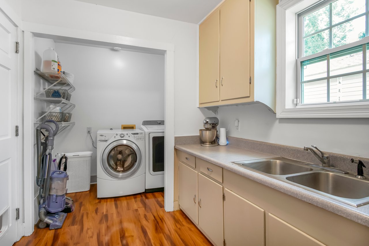 A bright laundry room with wooden floors, a washer and dryer, vacuum cleaner, beige cabinets, dual sinks, and a window letting in natural light.