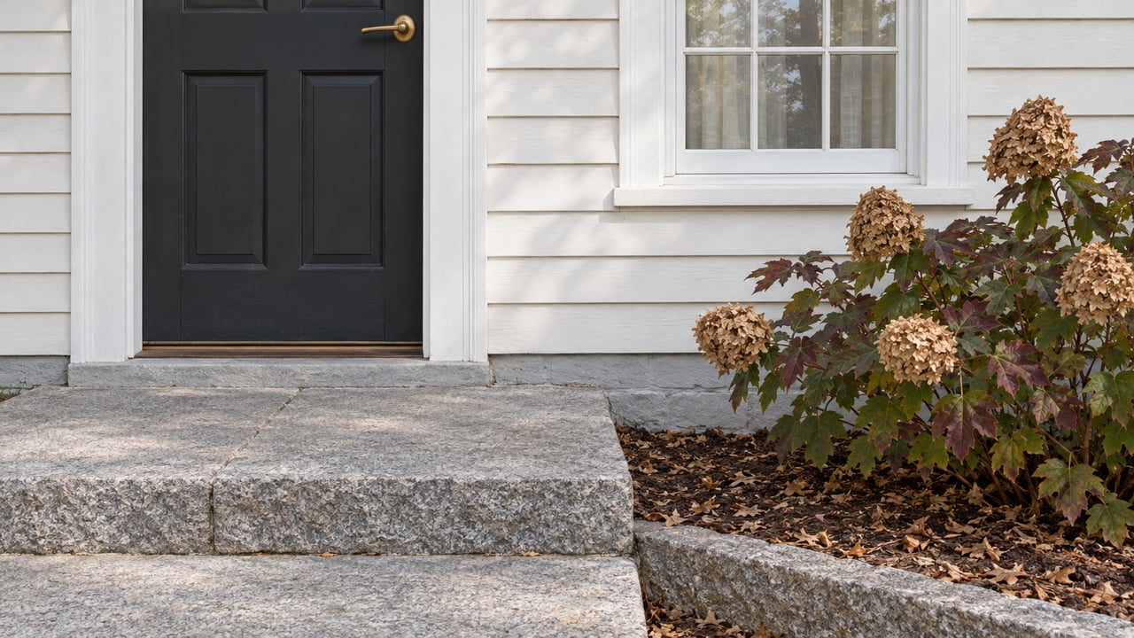 Close view of a white clapboard entry with a charcoal door, multi-pane window, worn granite landing and autumn oakleaf hydrangea.