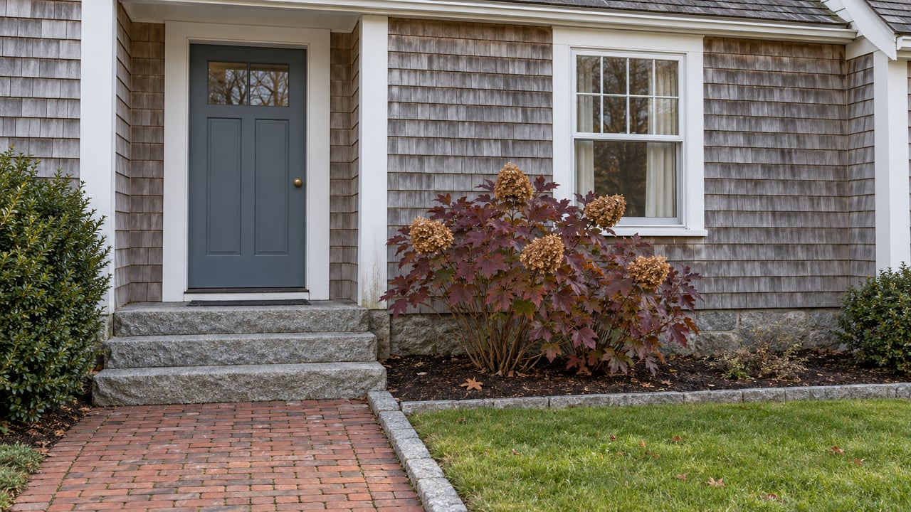 Weathered cedar-shingle home with a blue-gray entry door, granite steps, brick walk, and oakleaf hydrangea in late-autumn color.