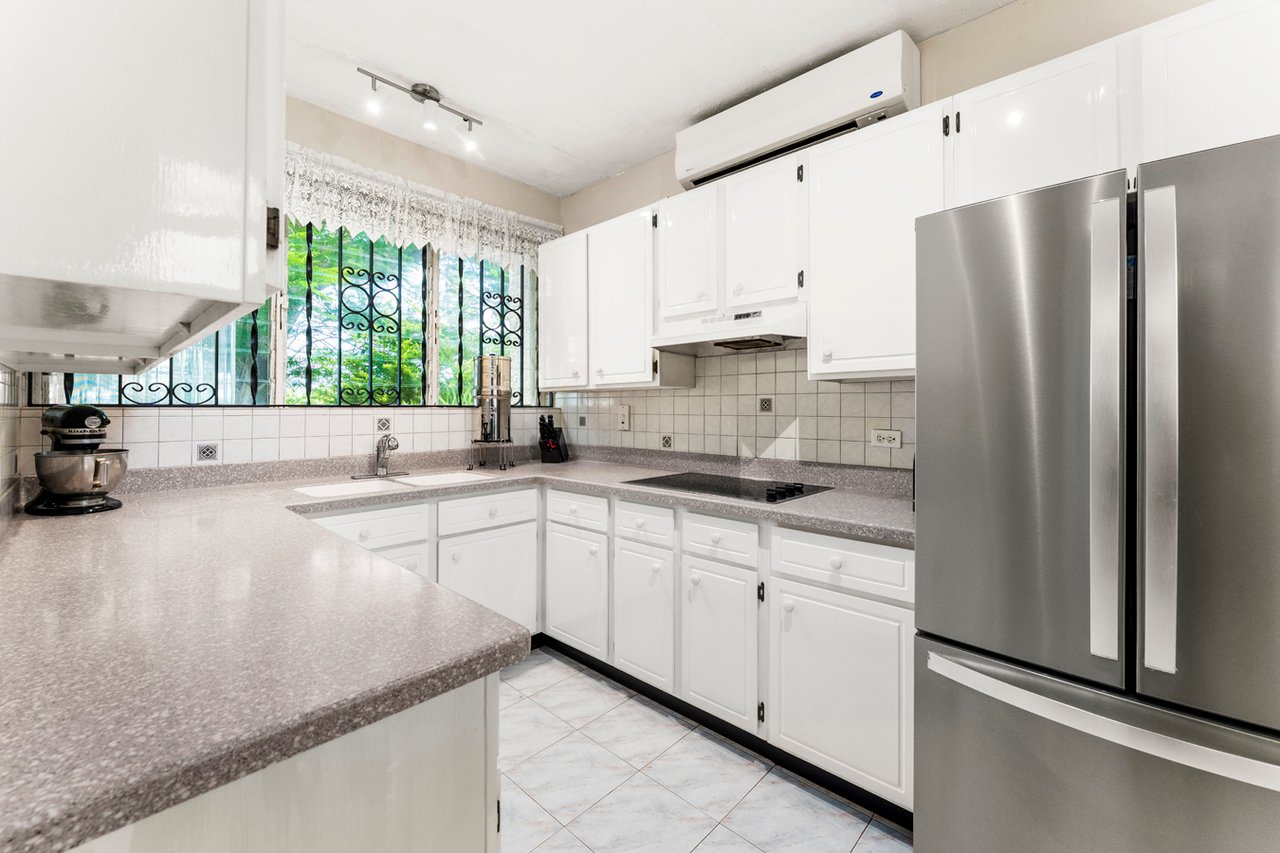 Kitchen with white cabinetry, speckled countertops, a stainless steel refrigerator, and a barred window overlooking greenery.
