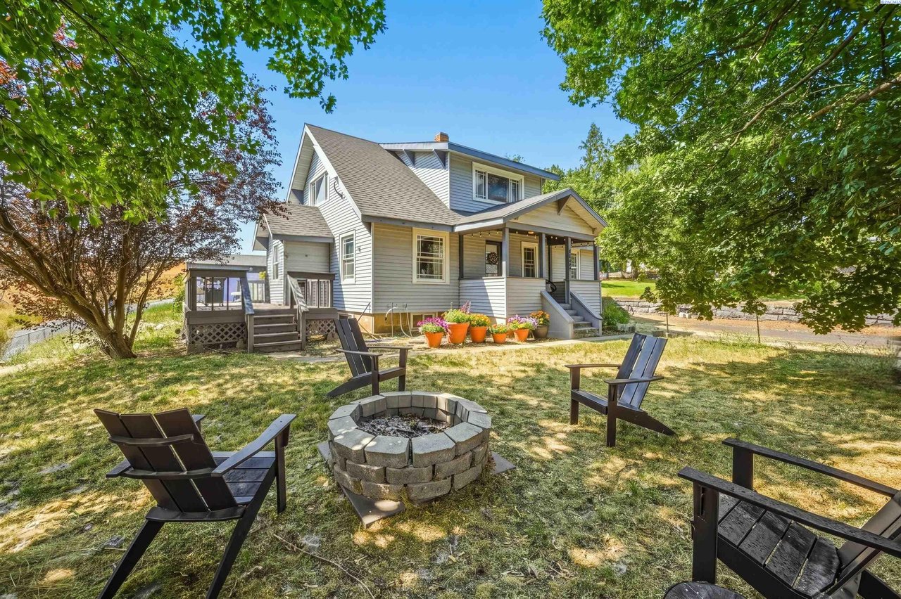 White two-story house with a covered porch, shaded lawn, Adirondack chairs, and a stone fire pit.