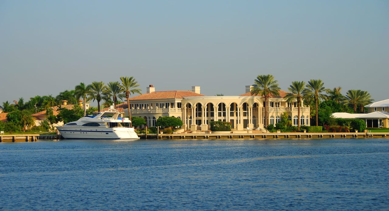 a large mansion is located in a tropical setting with palm trees and blue water with a yacht docked in front of it