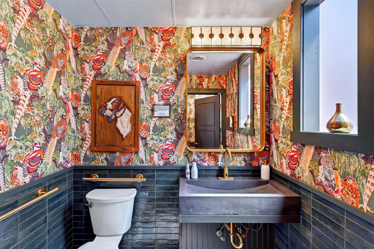 Restaurant restroom with colorful bird-patterned wallpaper, dark tile, a wall-mounted sink, and a large mirror.