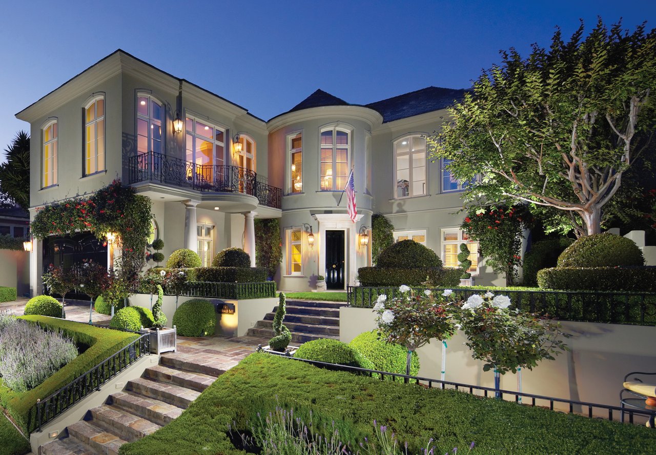 Two-story home with a curved balcony, illuminated windows, and formal gardens at dusk.