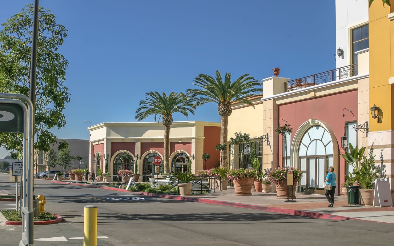 Commercial plaza with pastel storefronts, arched windows, palm trees, and flower planters.