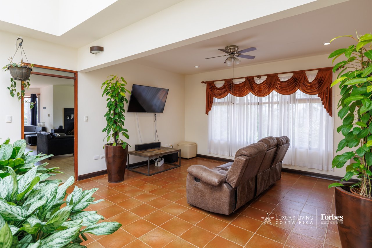 Living room with a brown reclining sofa, terracotta tile floors, potted plants, and a large curtained window.