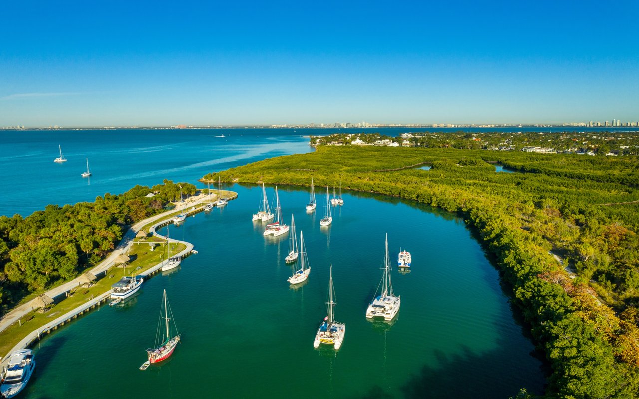 Aerial marina with moored sailboats, a green shoreline, and blue ocean beyond.