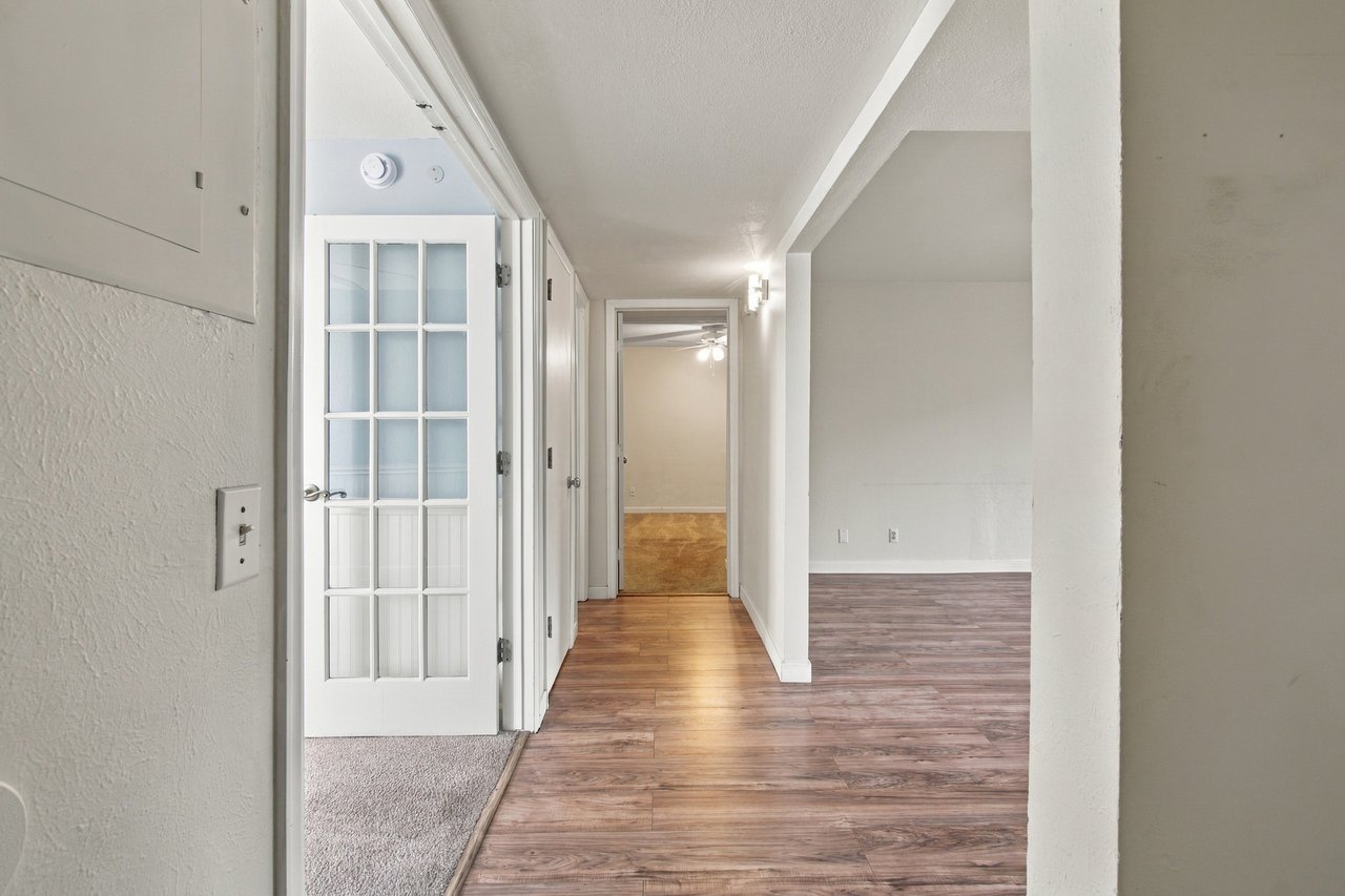 Hallway with wood-look flooring, a glass-paneled door, and an adjoining empty living area.