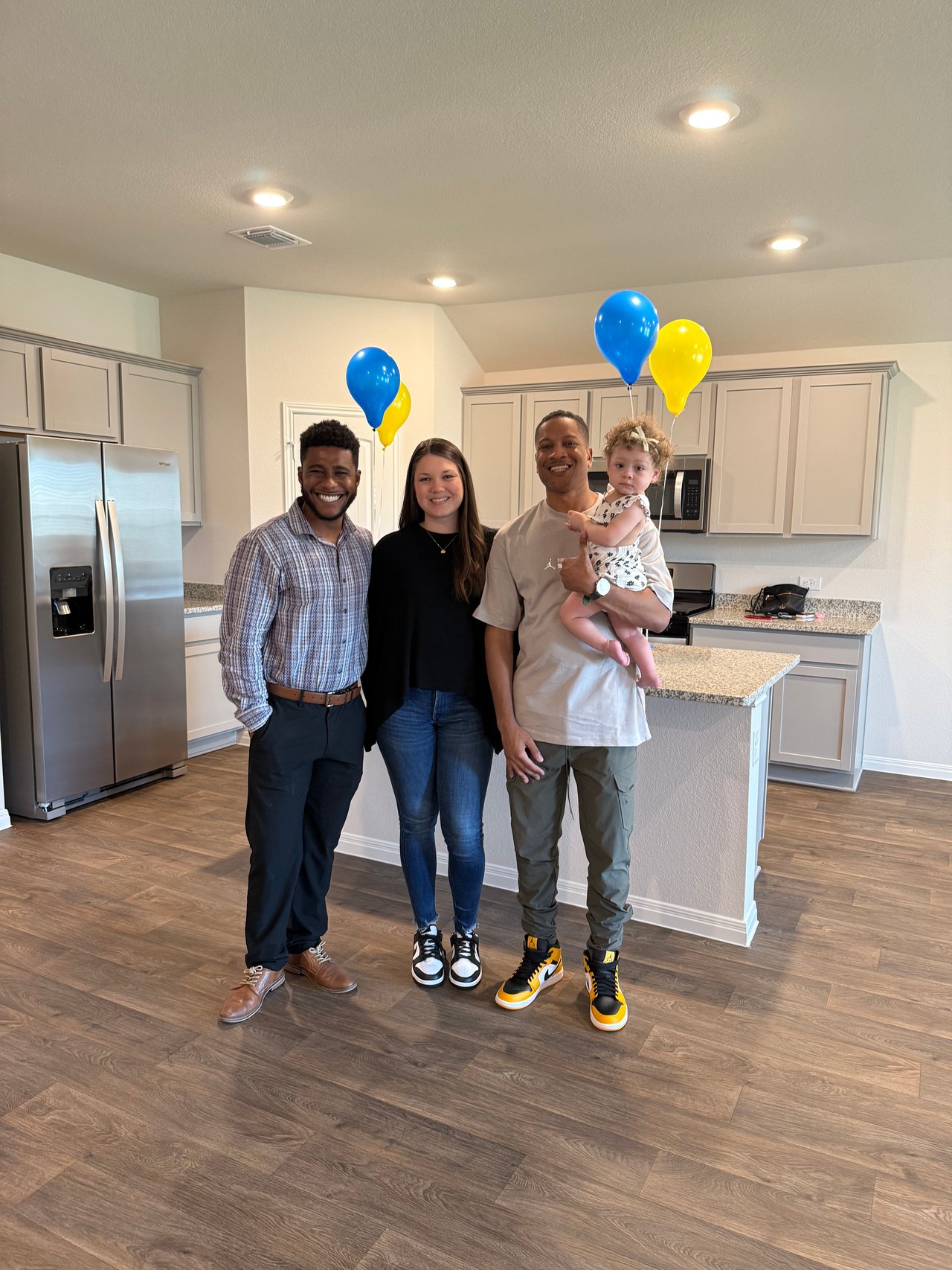 Three adults and a toddler pose in a bright kitchen with blue and yellow balloons.