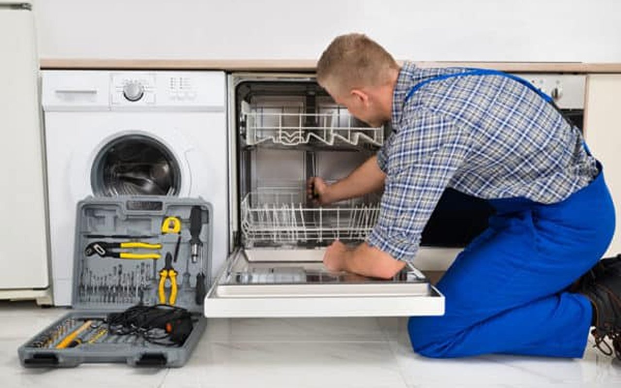 Repair technician works on an open dishwasher beside a washing machine and tool kit.