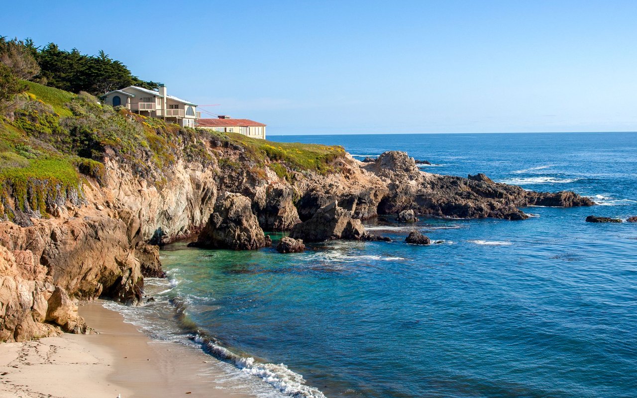 Coastal homes overlook a rocky bluff, Pacific Ocean, and small sandy beach.
