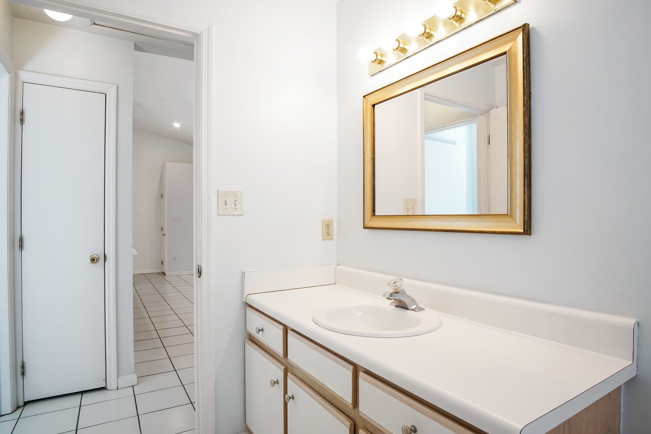 A bright bathroom with white walls and tiles features a countertop sink with cabinets below. A large mirror with a gold frame is above, reflecting light from five bulbs.