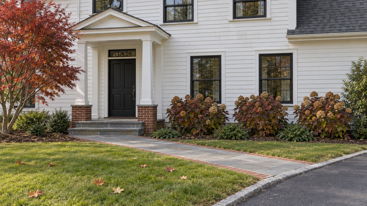 White clapboard home with black windows, a bluestone entry walk and autumn oakleaf hydrangeas beside a broad green lawn.