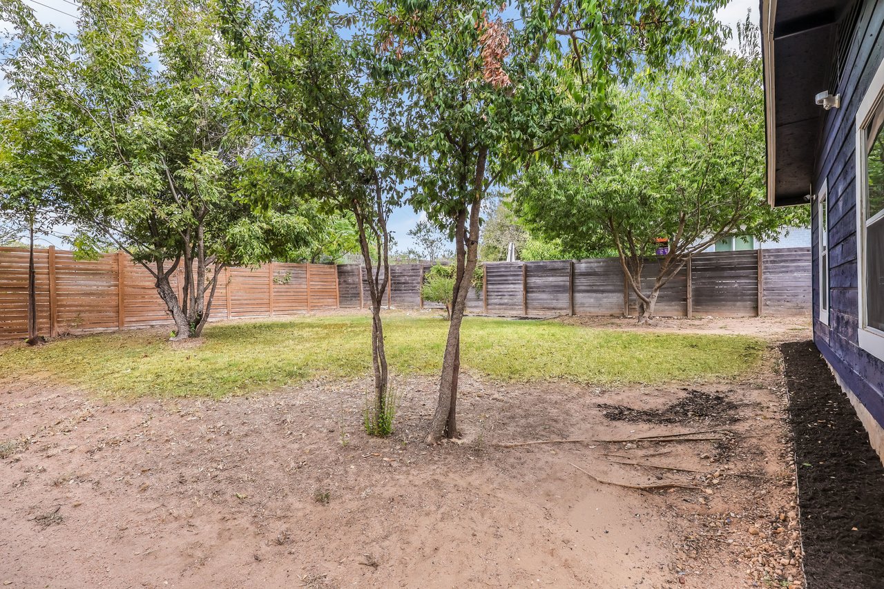 Fenced backyard with several leafy trees, patchy grass, and the house along one side.