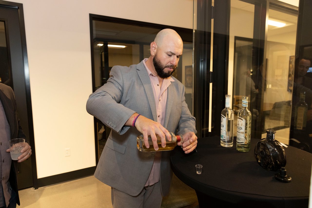 A man in a gray suit pours a drink at a table lined with liquor bottles.