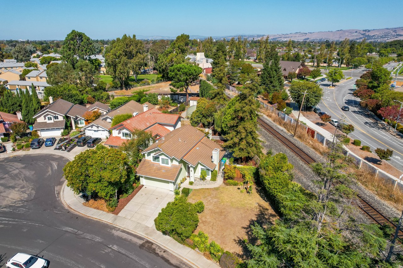 Aerial view of a corner home beside railroad tracks, with distant hills.