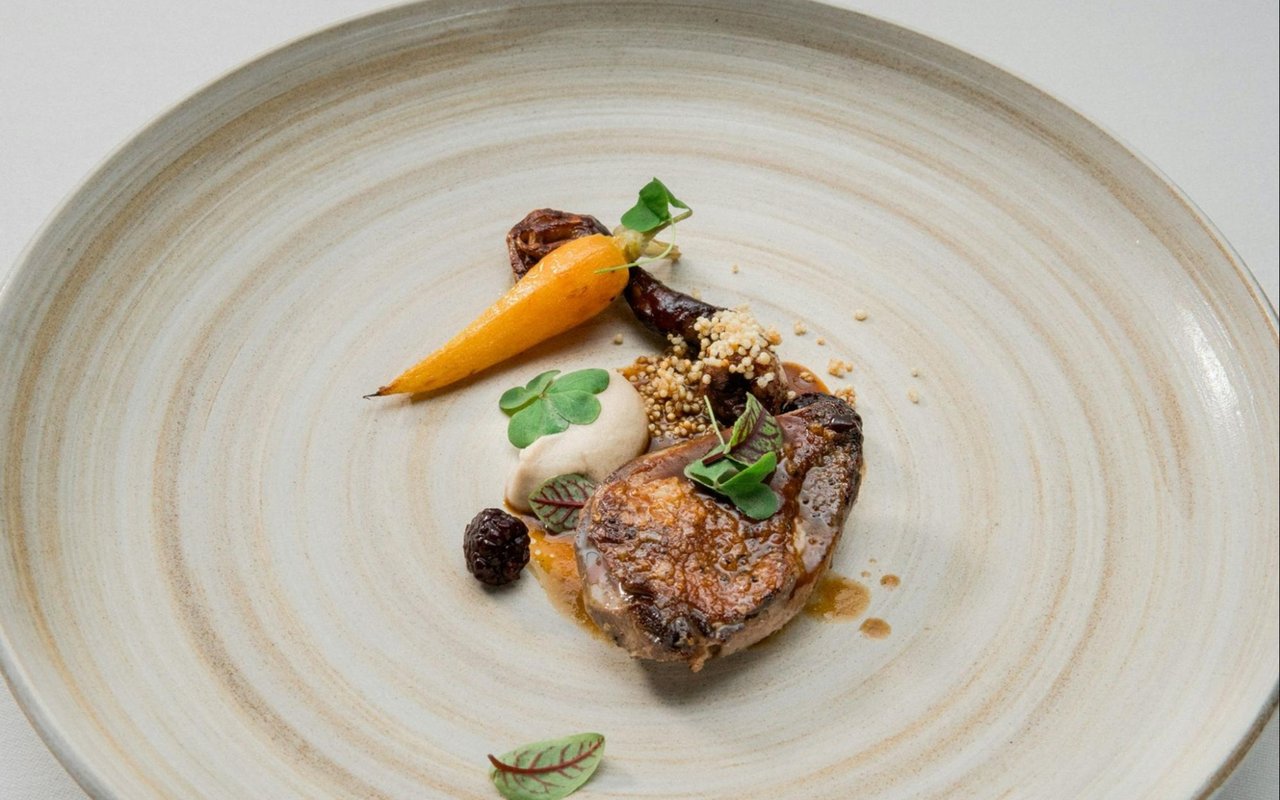 Seared meat with a roasted carrot, creamy purée, grains, and greens on a pale plate.