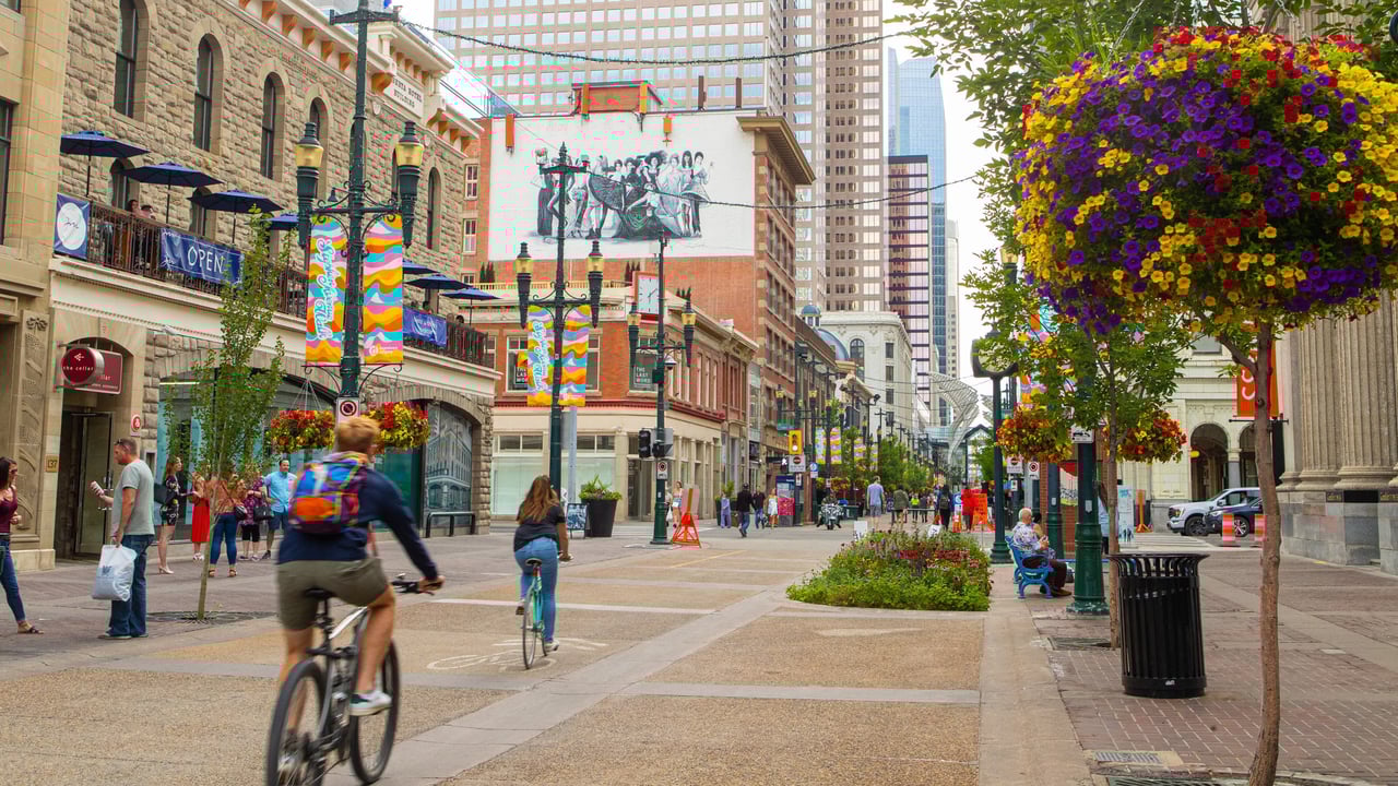 A lively summer day on Stephen Avenue with cyclists, shoppers, patios, and colorful hanging baskets brightening the downtown core.