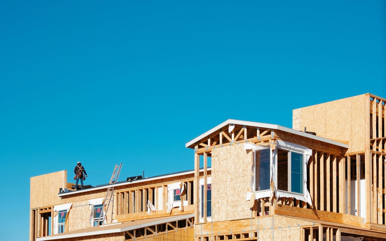 House under construction with exposed wood framing, a worker on the roof, and a ladder.