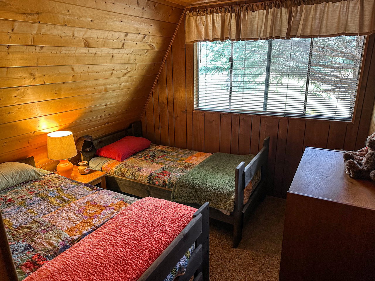 Bedroom with two single beds, colorful quilts, wood-paneled walls, and a window overlooking snowy trees.