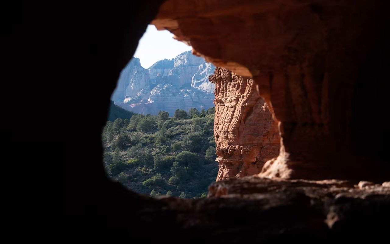 Mountain landscape and forested valley framed by a red sandstone cave opening.