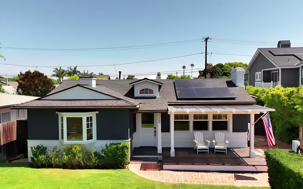  A dark blue single-story home with solar panels on the roof, a white porch, and a lush green lawn.