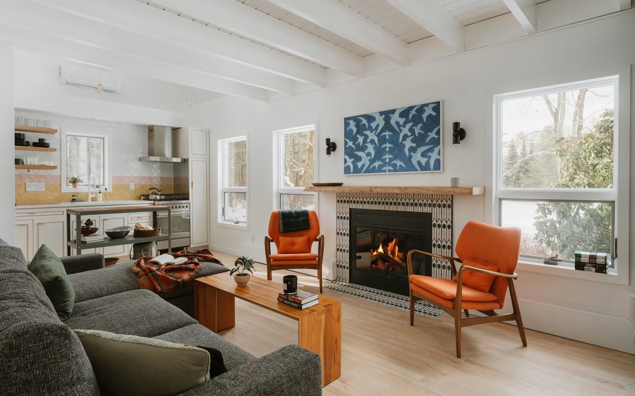 Living room with a lit patterned-tile fireplace, orange armchairs, exposed beams, and an open kitchen.