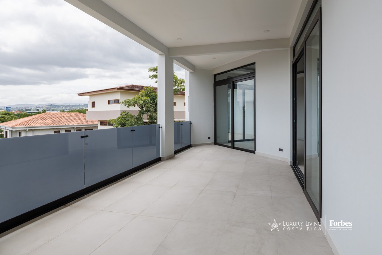 Covered apartment balcony with tiled flooring, glass railing, and views of neighboring homes and the distant city.