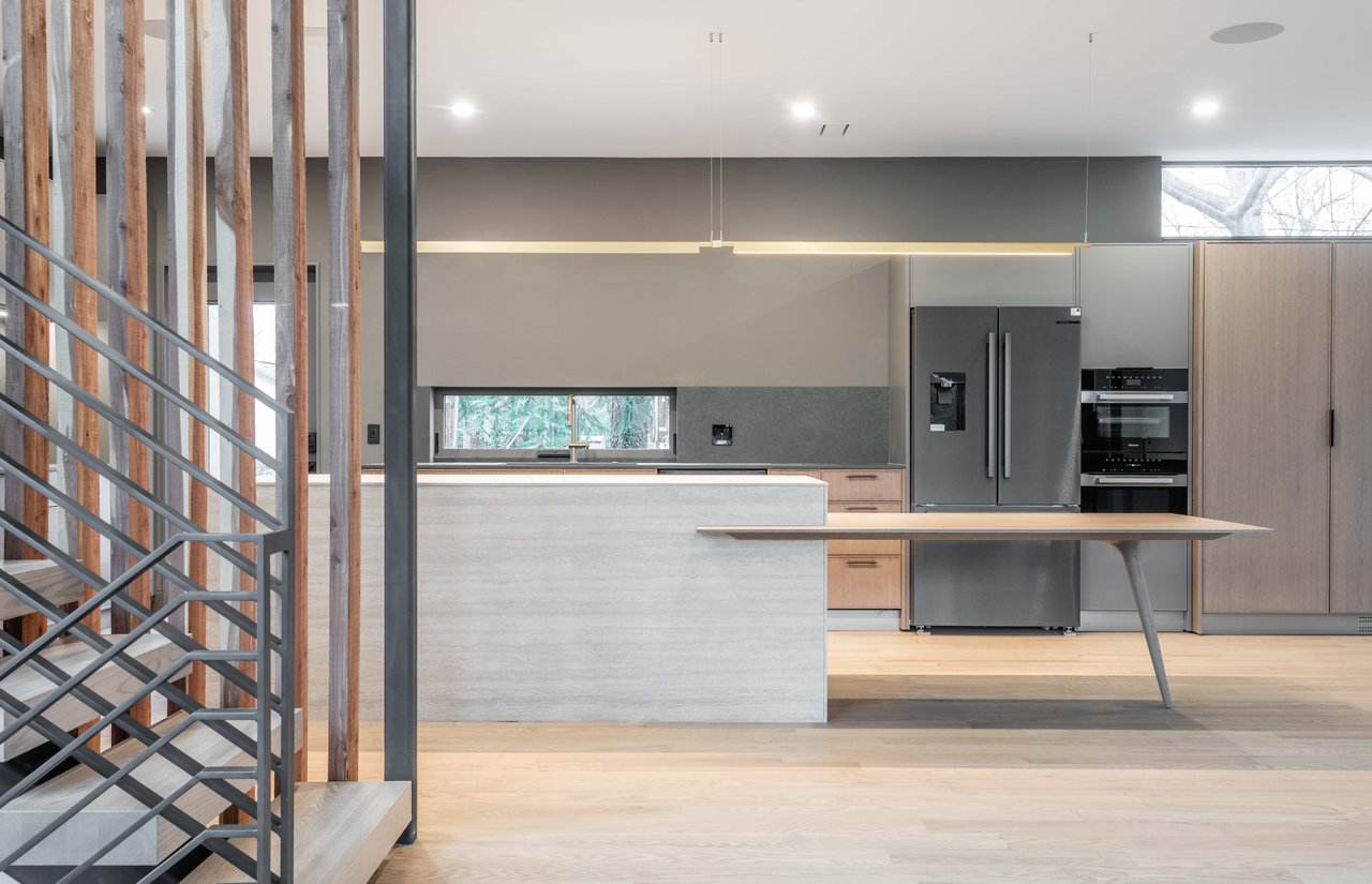 Modern kitchen with a large stone island, dark refrigerator, and metal staircase behind vertical wood slats.