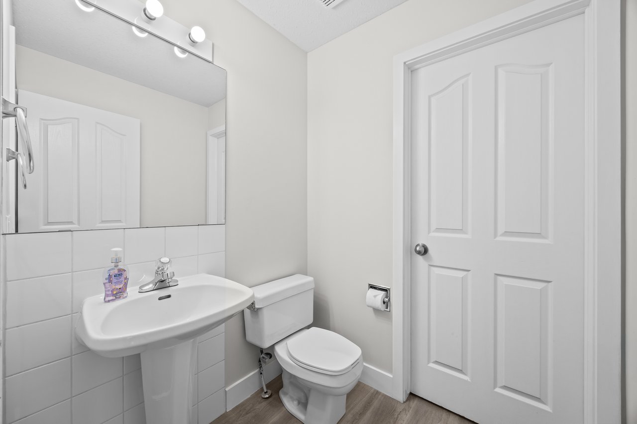 Compact bathroom with a pedestal sink, tiled backsplash, mirror, and toilet beside a white paneled door.