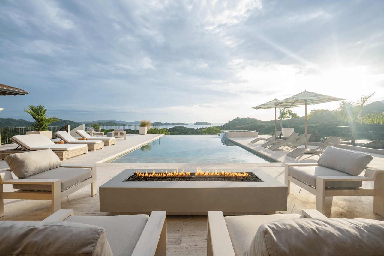 Infinity pool terrace with loungers and fire feature overlooking green hills and ocean.