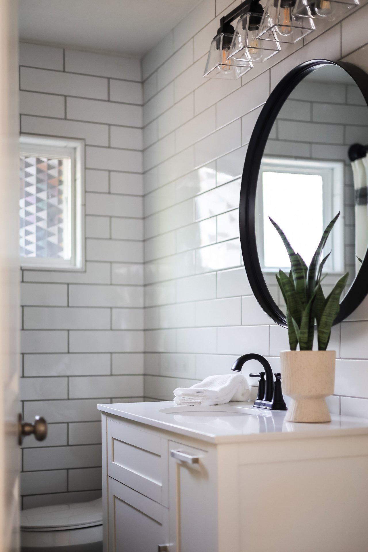 Bathroom vanity with black faucet, round mirror, white subway tile, and a potted snake plant.