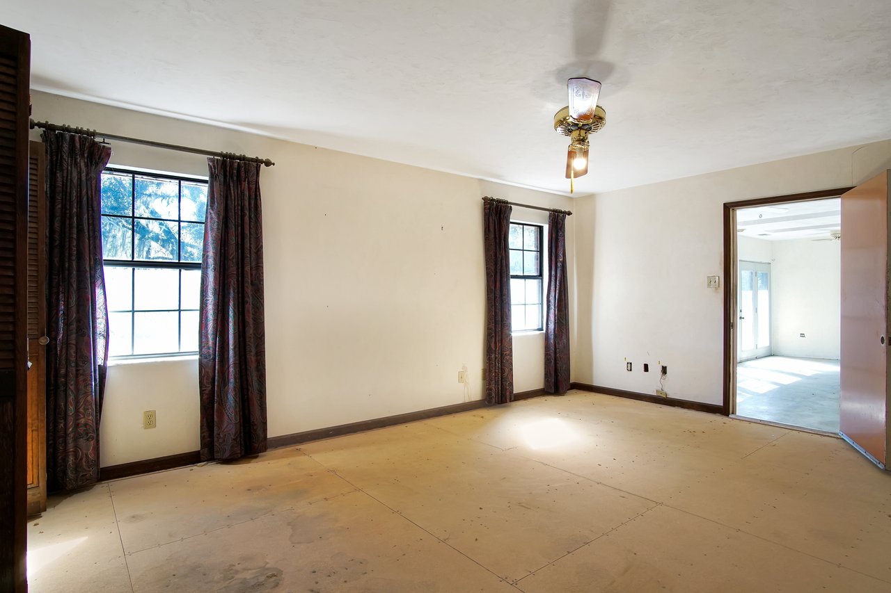 An empty room with bare floors, two curtained windows, and a ceiling fan. A doorway opens to a sunlit adjacent room, suggesting renovation.