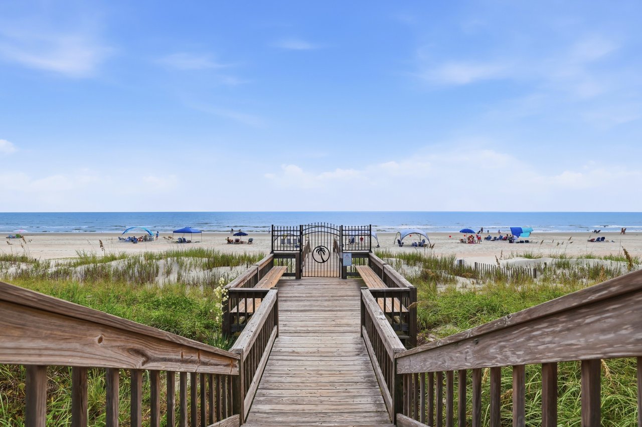 Beach-access boardwalk leads through grassy dunes to a sandy shore and ocean.