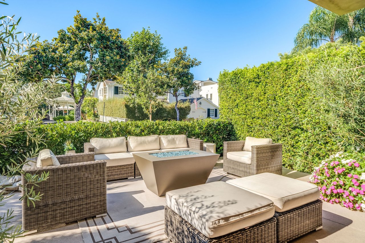 Furnished garden patio with wicker seating, cream cushions, and a glass-topped table surrounded by tall hedges.
