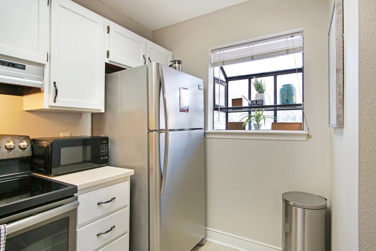 Compact kitchen with stainless steel fridge, microwave, and stove. A window with plants and a "Love Jesus" sign adds a cozy touch. Trash can nearby.