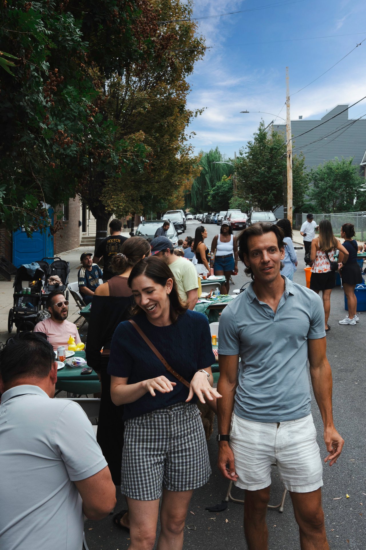 Neighbors mingle at a residential street block party; a smiling woman and man stand in the foreground.