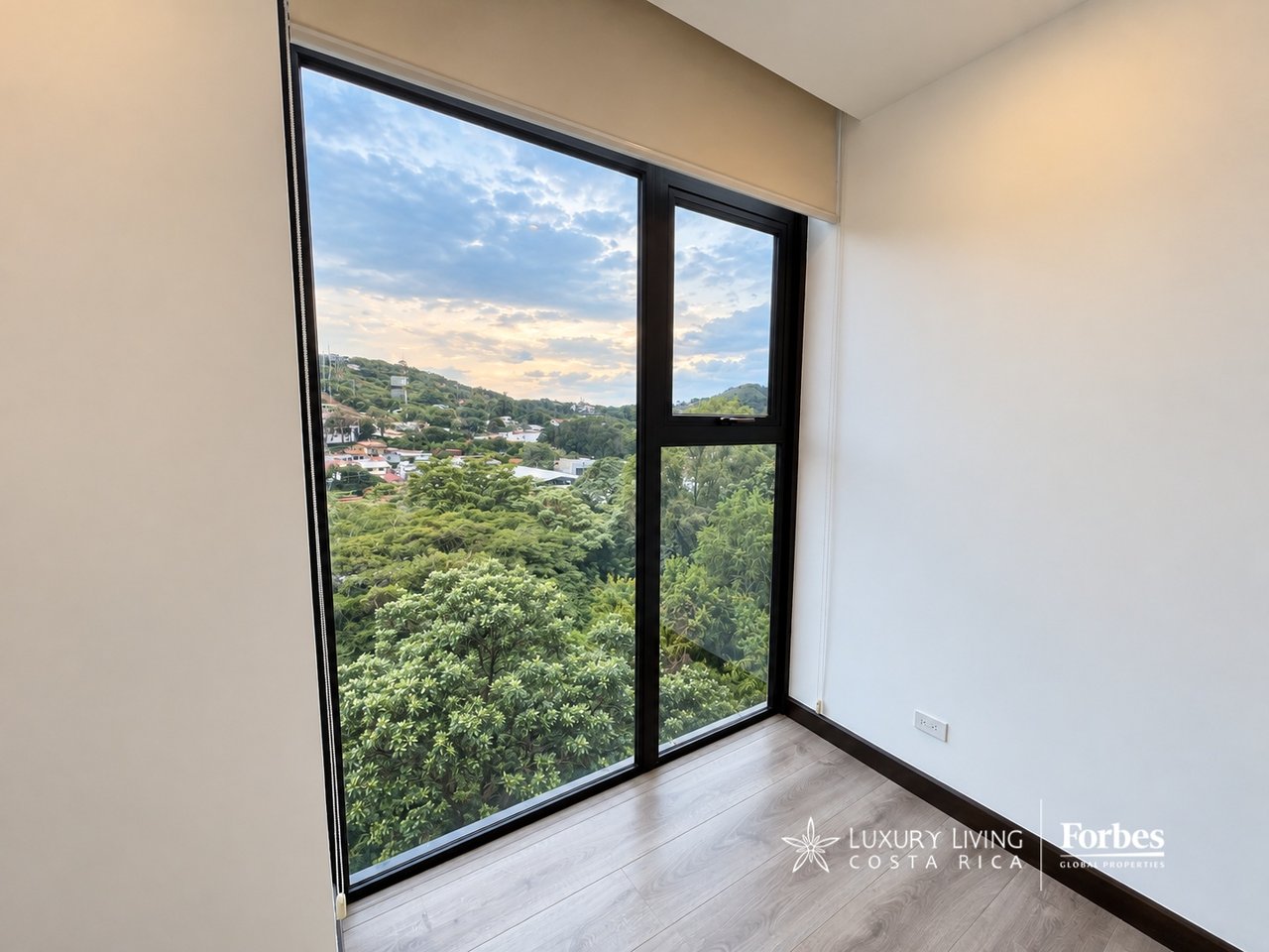 Empty apartment room with large windows overlooking leafy hills and homes.