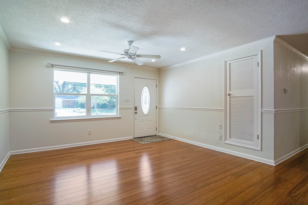 A bright, empty living room with wooden floors, a large window, white walls, and a ceiling fan. The front door has an oval glass design, creating a welcoming atmosphere.
