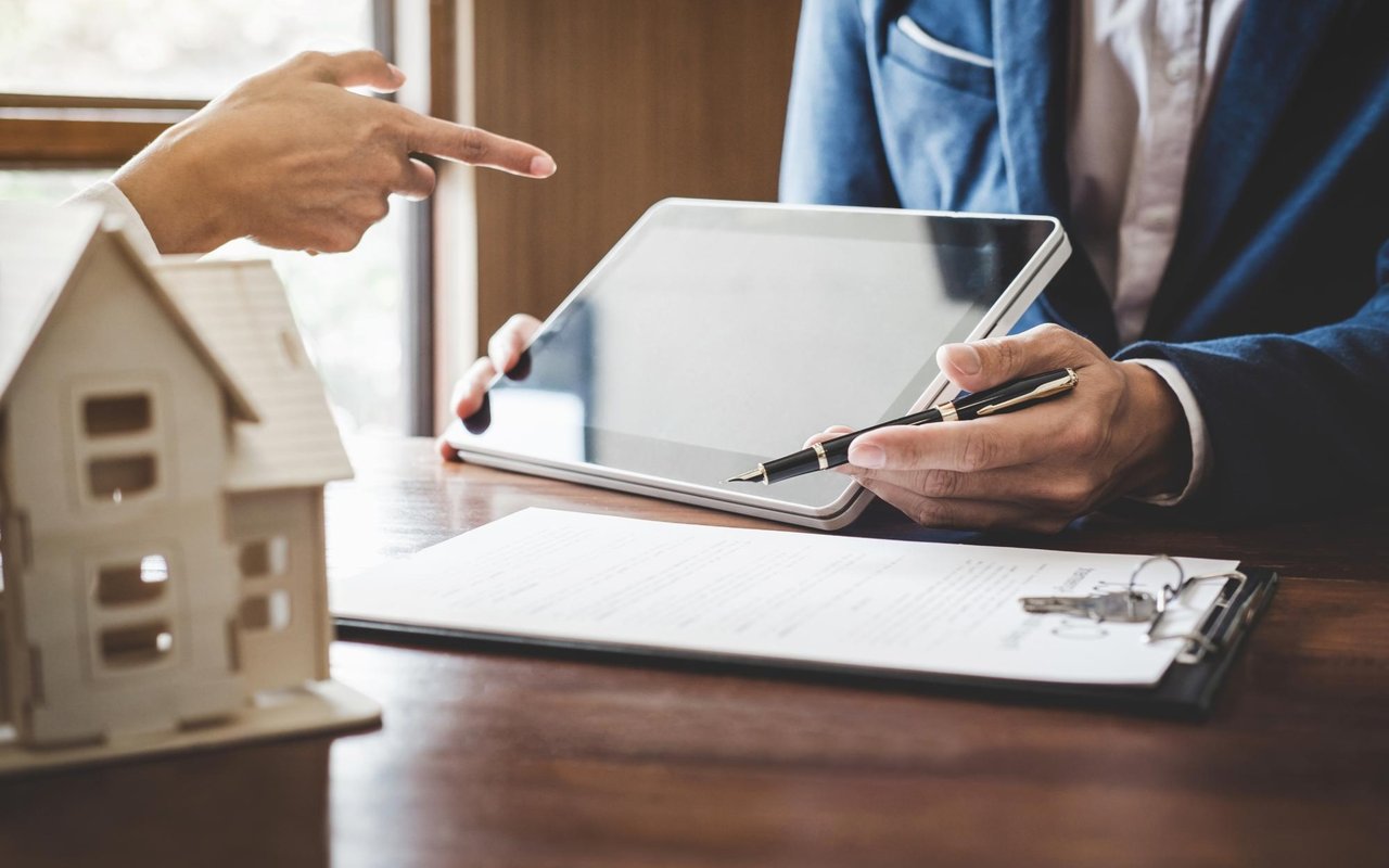 People review real estate paperwork on a tablet beside a model house and keys.