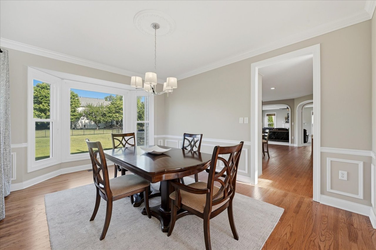 Dining room with bay windows overlooking a green backyard, hardwood floors, and white wainscoting.