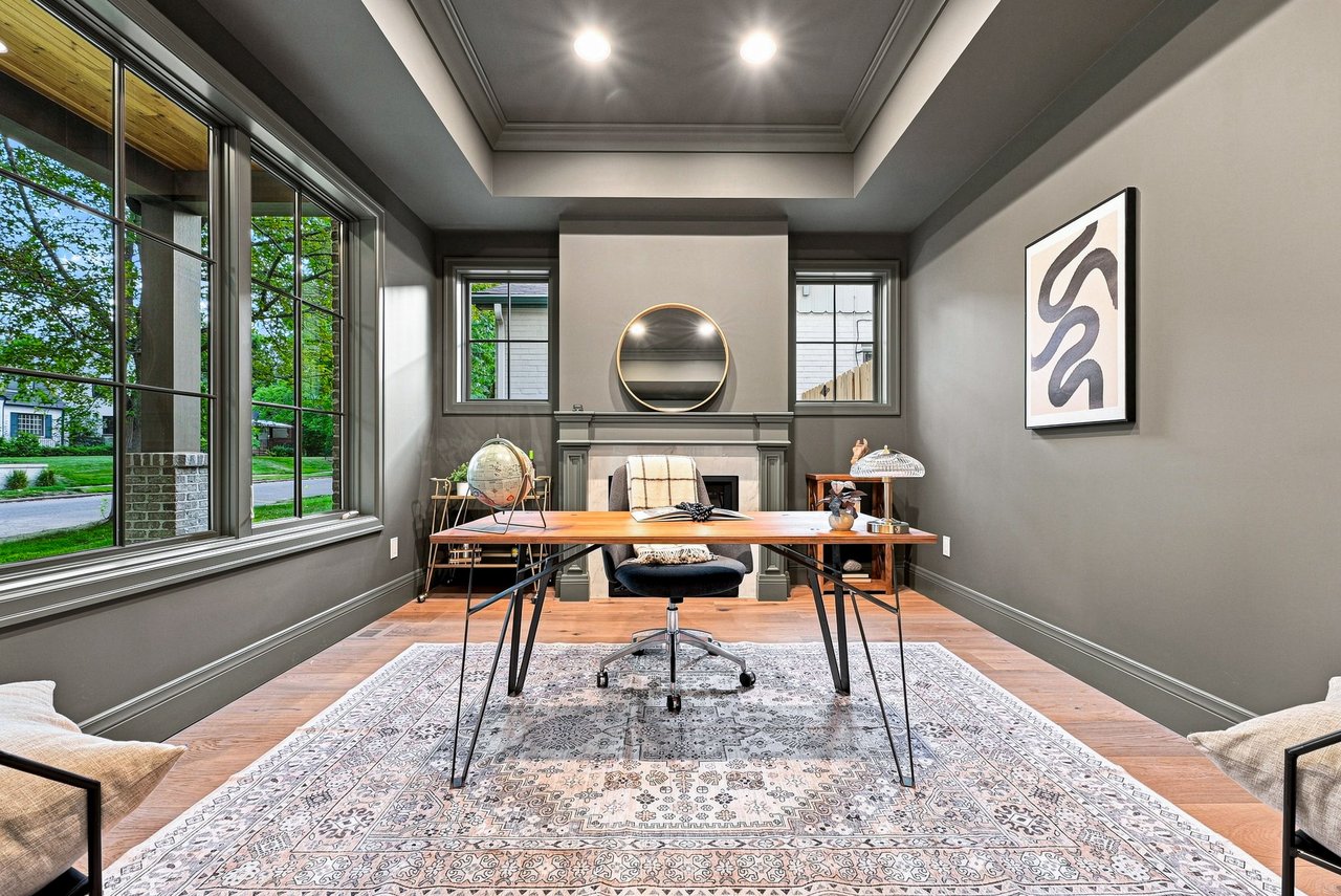 Furnished home office with a wooden desk, fireplace, round mirror, and large windows.