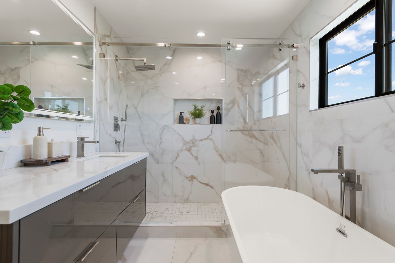 Marble bathroom with glass-enclosed shower, freestanding tub, and black-framed window.